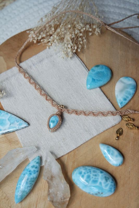Handgeknüpfter Choker mit tropfenförmigem Larimar-Anhänger, zartem Makramee-Band in Naturfarbe und kleinen Messingperlen. Schmuckstück liegt dekorativ auf Leinentuch mit getrockneten Blumen und Larimar-Rohsteinen.