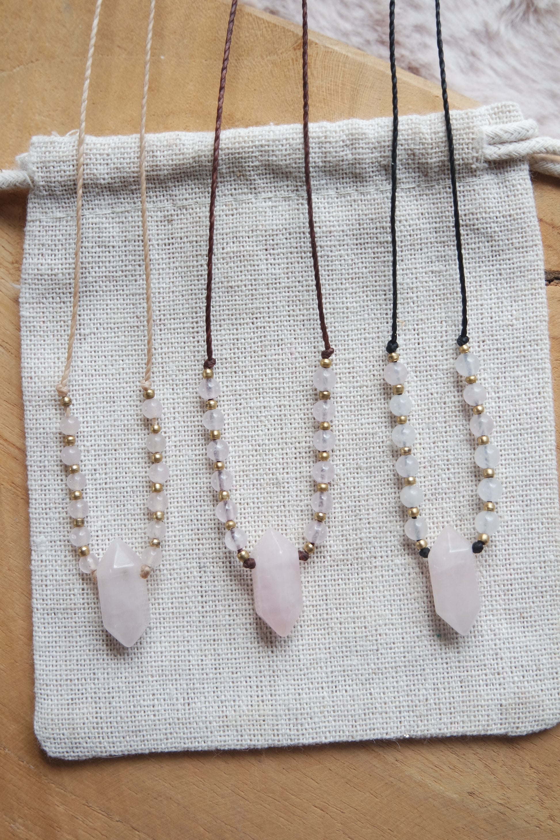 „Drei handgemachte Rosenquarz-Ketten in Braun, Schwarz und Beige auf naturfarbenem Leinenbeutel arrangiert.“