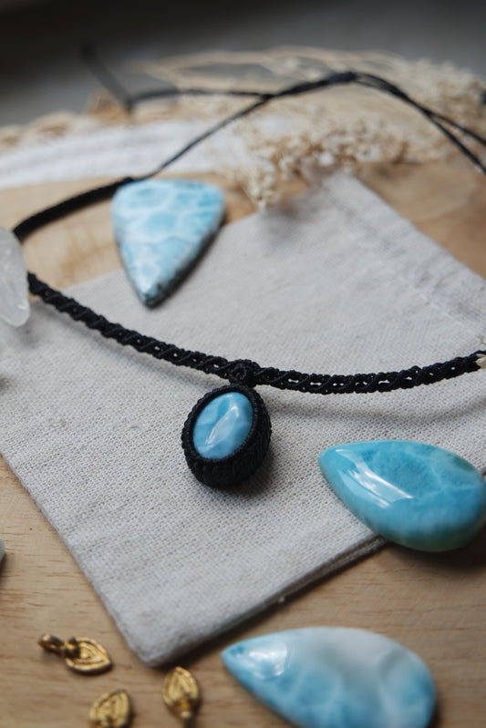 Schwarzer Makramee-Choker mit ovalem Larimar-Anhänger auf Leinentuch arrangiert, umgeben von Larimar-Rohsteinen.